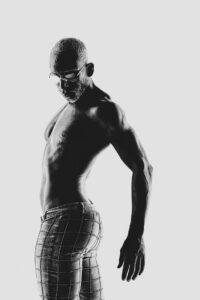 Shooting de David Dickens par Yan Forhan. Portrait en noir et blanc d’un homme torse nu portant des lunettes et un pantalon ajusté à motifs quadrillés.