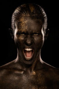 Portrait intense de Lorie, le regard perçant et la bouche entrouverte dans un cri, recouverte de peinture noire et d’éclats d’or. Photographie signée Yan Forhan, photographe officiel de Lorie Pester, avec le maquillage de Damien Dufresne. 4ᵉ de couverture de son livre photo.
