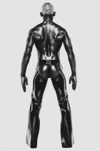 Shooting de David Dickens par Yan Forhan. Photographie en noir et blanc d’un homme vu de dos, torse nu, portant un pantalon en latex brillant avec un laçage ouvert à l’arrière.